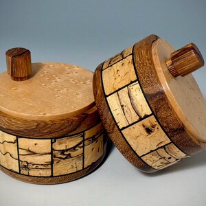 May include: Two cylindrical wooden containers with lids, each featuring a small knob handle. The sides are constructed from alternating segments of light-colored burl wood and dark brown wood, separated by thin black lines, with light-colored lids.