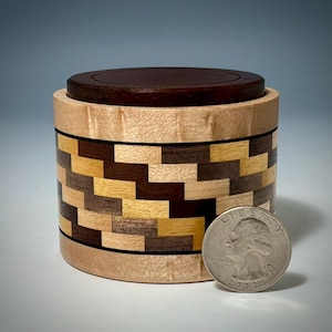 May include: A cylindrical wooden box with a dark brown lid and a body featuring a mosaic pattern of light, medium, and dark wood blocks. A United States quarter is placed next to the box to provide a visual reference for its scale.
