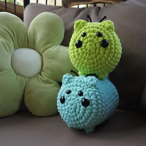 May include: Two crocheted plush toys in the shape of a cat. The top toy is green and the bottom toy is blue.