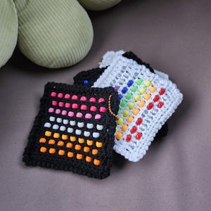 Può includere: Quattro quadrati all'uncinetto con perline colorate. I quadrati sono neri, bianchi e arcobaleno. I quadrati sono impilati uno sopra l'altro.