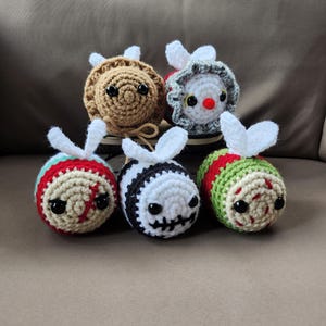 May include: Five crocheted bee plushies with different facial expressions. The bees are brown, white, red, green, and black. They have white wings and black eyes.