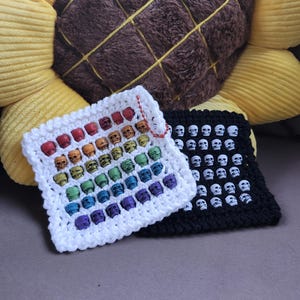 Könnte beinhalten: Zwei gehäkelte Quadrate, eines weiß und eines schwarz, sind mit Totenkopfperlen verziert. Das weiße Quadrat zeigt einen Regenbogenverlauf von Totenköpfen, während das schwarze Quadrat weiße Totenköpfe hat. Die Quadrate liegen auf einem Sonnenblumenkissen.