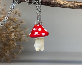 May include: A mushroom pendant necklace with a silver chain. The pendant features a red cap with white polka dots and a white stem. The necklace is hanging from a branch.