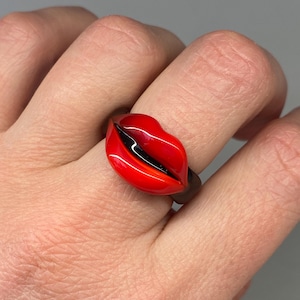 Puede incluir: Un llamativo anillo con forma de labios rojos, con un acabado brillante. El anillo presenta una banda oscura y una detallada representación de labios, con una línea negra que representa la abertura de la boca. Un accesorio único y llamativo.