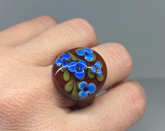 May include: A round ring with a brown base and a floral design. The ring features blue flowers with white accents and green leaves. The ring is a unique piece of jewellery.