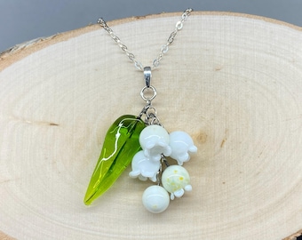 May include: A silver chain necklace with a pendant featuring a cluster of white glass lily of the valley flowers and a single green leaf.