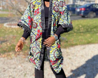 Op de afbeelding: Een patroonkimono met een zwarte satijnen rand. De kimono heeft een levendig ontwerp met rode, gele en groene accenten. Het model draagt een zwarte top en broek, en zwarte laarzen. De achtergrond is een zonnige buitenscène.