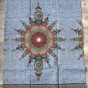 May include: A rectangular piece of fabric featuring a grey background with a dark grey cross-hatch pattern. A large, symmetrical, star-shaped medallion in red, green, and blue sits in the centre. The top and bottom borders feature black bands with colourful geometric and floral motifs, including the text "GUARANTEED REAL WAX BLACK MAMA".