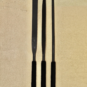 Peut inclure: Trois limes en métal noir avec des poignées en caoutchouc noir. Les limes sont de différentes longueurs et ont des formes différentes.