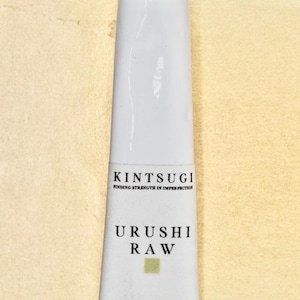 Op de afbeelding: Een witte tube Kintsugi Urushi Raw, een soort Japanse lak die wordt gebruikt om gebroken aardewerk te repareren. De tube heeft de tekst "Kintsugi Finding Strength in Imperfection" en "Urushi Raw" erop gedrukt. De Japanse karakters "金継ぎ" staan ook op de tube.