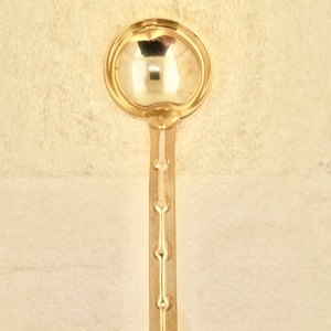 May include: A gold-colored ladle with a round bowl and a long handle. The handle has a decorative design of small circles.