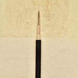 Peut inclure: Un outil d'art à pointe fine, probablement un stylo ou un pinceau, avec un design noir et beige, sur un fond beige clair texturé. L'outil a un manche fin et foncé et une pointe métallique, idéal pour les détails.