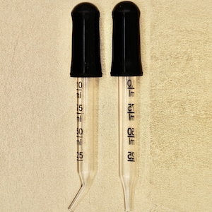 Könnte beinhalten: Zwei klare Glaspipetten mit schwarzen Gummikappen. Die Pipetten sind in Millilitern von 25 bis 75 gekennzeichnet.