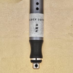 Puede incluir: Primer plano de una pluma negra y plateada con las palabras "Golden Joint" y caracteres japoneses impresos en el lateral.