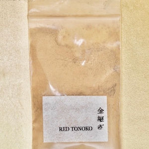 Może przedstawiać: Mały, przezroczysty plastikowy woreczek wypełniony jasnobrązowym proszkiem. Woreczek ma białą etykietę z tekstem "RED TONOKO" i japońskimi znakami, które tłumaczą się jako "Złoty proszek".