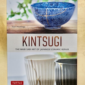 Może przedstawiać: Okładka książki przedstawiająca niebiesko-białą ceramiczną miskę ze złotymi akcentami, prezentująca japońską sztukę Kintsugi. Tytuł "Kintsugi" jest wyraźnie widoczny, z podtytułem "Sztuka Wabi Sabi naprawy japońskiej ceramiki."