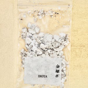 Peut inclure: Un sac en plastique transparent rempli de petits cailloux blancs et gris. Une étiquette blanche avec le mot "IBOTA" et des caractères japonais est attachée. Le fond est de couleur beige clair.