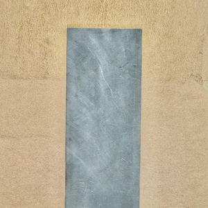 Puede incluir: Primer plano de una piedra rectangular gris con una superficie lisa. La piedra está colocada sobre un fondo beige claro.