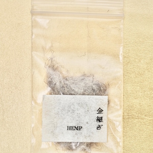 Może przedstawiać: Mały, przezroczysty woreczek z tworzywa sztucznego z białą etykietą, na której widnieje napis "HEMP" i "金継ぎ" w języku japońskim. Woreczek zawiera niewielką ilość szarych włókien konopnych.