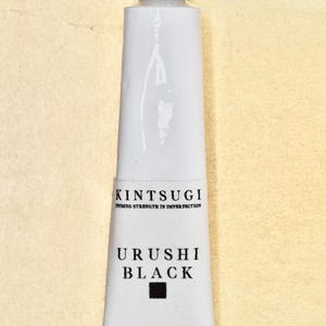 May include: A white tube of Kintsugi Urushi Black paint. The tube has the words "Kintsugi Finding Strength in Imperfection" and "Urushi Black" printed on it. The tube also has a black square and Japanese characters.