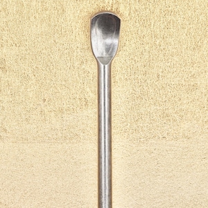 May include: A silver metal tool with a rounded, spoon-like tip. The tool is on a textured, light beige background.