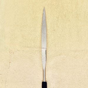 Op de afbeelding: Een zilveren metalen vijl met een zwarte handgreep. De vijl is scherp en puntig.