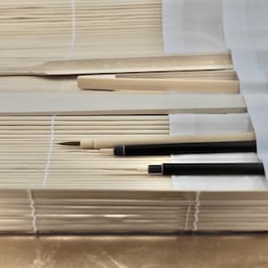 Puede incluir: Un conjunto de pinceles de caligrafía y una regla se colocan sobre una estera de bambú. Los pinceles están envueltos en un paño blanco y la regla es de madera.