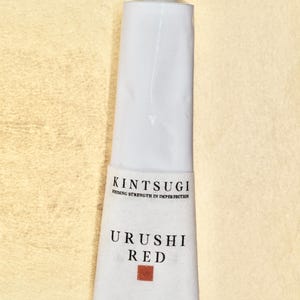 May include: A white tube of Kintsugi Urushi Red paint. The tube has the words "Kintsugi Finding Strength in Imperfection" and "Urushi Red" printed on it. The tube also has a small red square and the Japanese characters for "gold repair" printed on it.