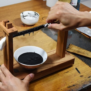 Peut inclure: Un support en bois pour l'encre avec une barre recouverte d'encre noire, laissant couler de l'encre dans un bol blanc. Une personne essuie l'encre avec un chiffon blanc. Un autre bol avec du liquide blanc et un outil en bois sont également visibles.