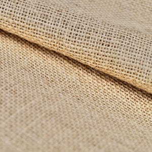 Op de afbeelding: Close-up van een natuurlijke bruine jute stof met een geweven textuur. De stof is dubbelgevouwen, waardoor de onderkant zichtbaar is.