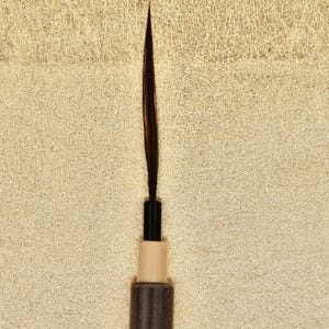 Peut inclure: Gros plan sur un pinceau eyeliner marron foncé avec un manche noir et une base beige clair. Le pinceau a une pointe fine et pointue, idéale pour une application précise. Fond beige clair texturé.