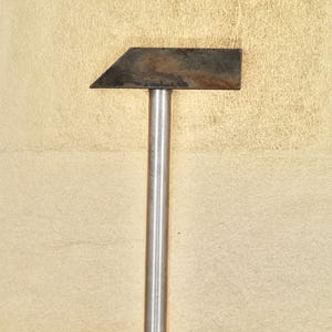 Op de afbeelding: Een zilverkleurige hamer met een rechthoekige kop. De kop is gemaakt van donker metaal en heeft een roestige uitstraling.