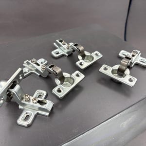 Puede incluir: Varias bisagras plateadas para armarios están dispuestas sobre una superficie gris. Cada bisagra tiene un brazo metálico curvado y una placa de montaje con orificios para tornillos. Diseñadas para puertas de armarios y muebles.