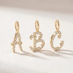 Op de afbeelding: Drie gouden letterhangers, A, B en C, met een gepolijste gouden afwerking en een reeks kleine, heldere, fonkelende steentjes ingelegd op de voorzijde van de letters, elk voorzien van een klein gouden oogje voor bevestiging.