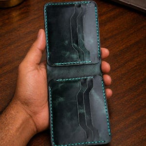 Può includere: Portafoglio in pelle verde scuro con cuciture turchese. Il portafoglio presenta più fessure per carte e un design a due ante. La pelle ha un aspetto invecchiato e le cuciture forniscono un accento a contrasto. Il portafoglio è tenuto aperto, mostrando l'interno.