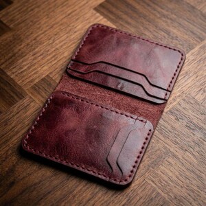 Può includere: Portafoglio in pelle bordeaux aperto con più fessure per carte. Il portafoglio è aperto, rivelando le fessure interne per carte. La pelle ha un aspetto invecchiato, con venature e variazioni di colore visibili. I bordi sono cuciti con un filo a contrasto.