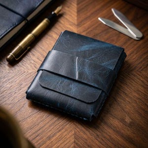 Puede incluir: Cartera de cuero azul oscuro con cierre de correa, sobre una superficie de madera. También se ven una pluma estilográfica de latón, un cuaderno y un clip de dinero plateado. La cartera tiene un aspecto texturizado.