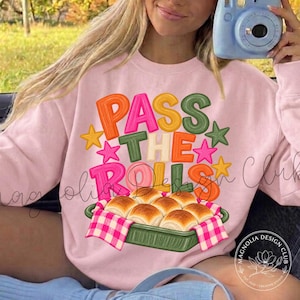 May include: A light pink sweatshirt featuring the text "PASS THE ROLLS" in colorful, bubbly letters surrounded by small stars. Below the text is a graphic of a green baking dish filled with golden-brown dinner rolls on a pink checkered cloth.