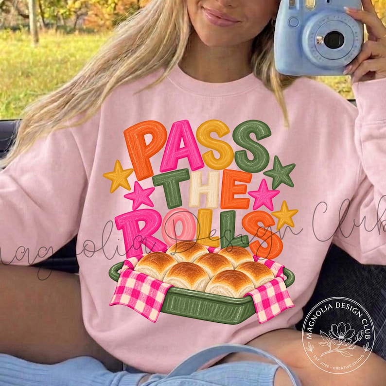 May include: A light pink sweatshirt featuring the text "PASS THE ROLLS" in colorful, bubbly letters surrounded by small stars. Below the text is a graphic of a green baking dish filled with golden-brown dinner rolls on a pink checkered cloth.