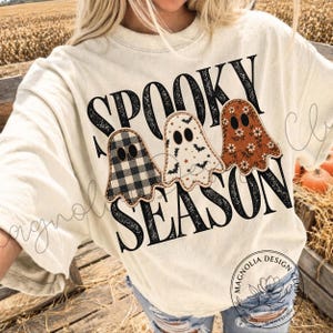 May include: A cream-coloured t-shirt featuring the black text SPOOKY SEASON and three ghost illustrations. The first ghost has a black and white buffalo check pattern, the second has a white background with bats, and the third has a rust floral print.