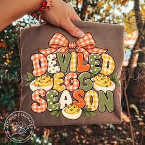 May include: A brown t-shirt featuring a large graphic with the text "DEVILED EGG SEASON" in colorful, patterned letters. The design includes a large orange and white gingham bow at the top and three illustrations of deviled eggs.