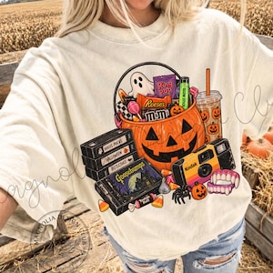 Puede incluir: Una camiseta color crema con un estampado gráfico de un cubo de calabaza lleno de dulces, una pila de cintas VHS etiquetadas como Hocus Pocus, Halloween, Beetlejuice y Goosebumps, una cámara Kodak, un vaso de bebida y dientes de vampiro.