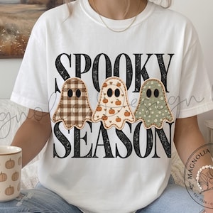 Puede incluir: Una camiseta blanca con el texto negro SPOOKY SEASON y tres ilustraciones de fantasmas. El primer fantasma tiene un patrón de cuadros marrones, el segundo es crema con calabazas naranjas y el tercero es verde salvia con un pequeño patrón floral.
