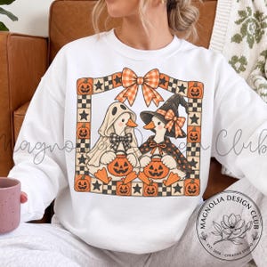 May include: A white crewneck sweatshirt featuring a graphic of two ducks dressed for Halloween, one as a ghost and one as a witch, framed by a border of orange pumpkins, black stars, and checkered patterns with a large orange plaid bow on top.