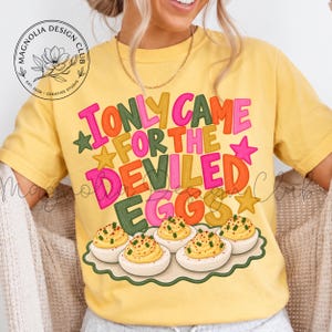Puede incluir: Una camiseta amarilla de manga corta con el texto I ONLY CAME FOR THE DEVILED EGGS en letras coloridas y llamativas. Debajo del texto hay una ilustración de cinco huevos rellenos en una bandeja de borde festoneado. Un logo está arriba a la izquierda.
