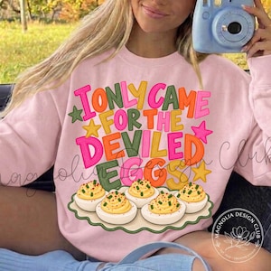 May include: A light pink crewneck sweatshirt featuring a colorful graphic print with the text I ONLY CAME FOR THE DEVILED EGGS in pink, yellow, orange, and green letters, accompanied by a platter of six deviled eggs garnished with green herbs.