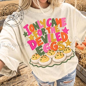 May include: A cream-colored t-shirt featuring a graphic print with the text I ONLY CAME FOR THE DEVILED EGGS in colorful block letters. The design includes small stars and a platter of five deviled eggs topped with green herbs and red paprika.