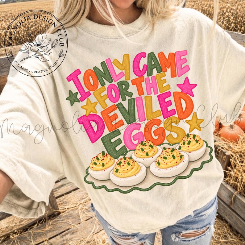 May include: A cream-colored t-shirt featuring a graphic print with the text I ONLY CAME FOR THE DEVILED EGGS in colorful block letters. The design includes small stars and a platter of five deviled eggs topped with green herbs and red paprika.