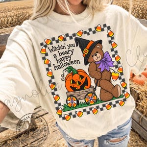 Peut inclure: Un t-shirt couleur crème avec un motif d'ours en peluche marron portant un chapeau de sorcière noir et un nœud violet. L'ours est à côté d'un chariot en bois contenant une citrouille d'Halloween. Le texte indique Witchin' you a beary happy halloween.