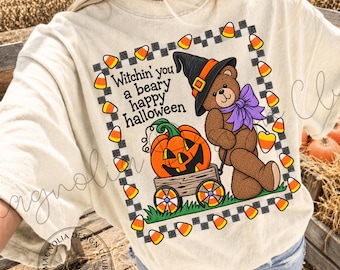 May include: A cream-colored t-shirt featuring a graphic of a brown teddy bear wearing a black witch hat and a purple bow. The bear stands next to a wooden wagon containing an orange jack-o'-lantern. The text reads Witchin' you a beary happy halloween.